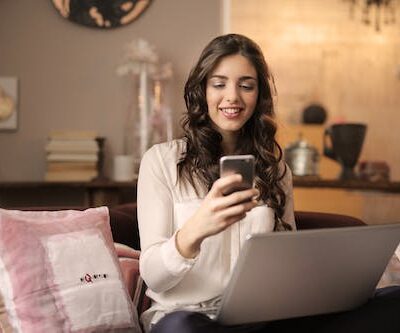 Free Woman Sitting on Sofa While Looking at Phone With Laptop on Lap Stock Photo